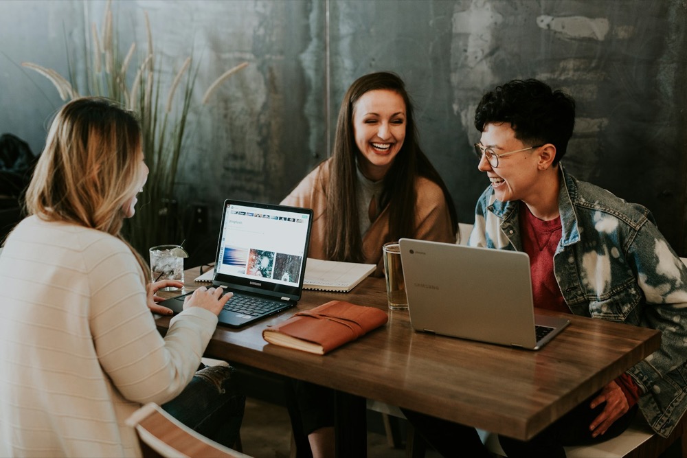 Drie mensen lachen samen achter laptops aan een werktafel